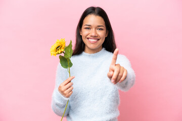 Young Colombian woman holding sunflower isolated on pink background showing and lifting a finger