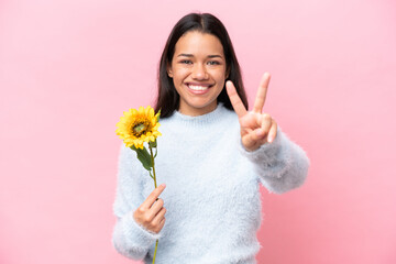 Young Colombian woman holding sunflower isolated on pink background smiling and showing victory sign