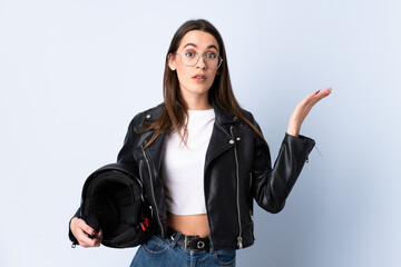 Young woman holding a motorcycle helmet over isolated blue background with surprise facial...