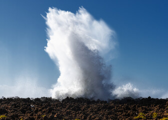 wave crashing onto a rocky reef under a blue sky and creating an eruption of water