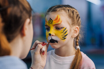 A woman professional artist paints with a brush, a colored pencil on the face of a child, a beautiful girl, face painting, makeup, drawing at a children's party. Photography, creative process.