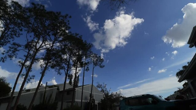 Low angle timelapse of sunny blue sky with white clouds over trees and buildings with parked car