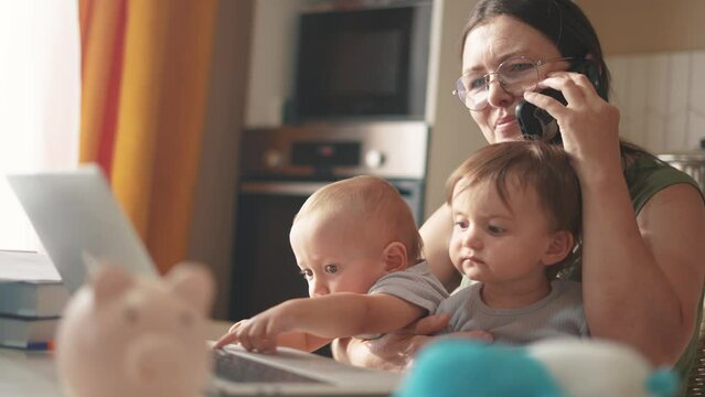 mother works remotely from home with two babies in her arms. pandemic remote work concept. mom tries to work at home in a fun kitchen, small children interfere with sitting in business her arms