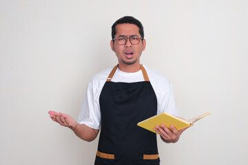 An Asian man wearing apron showing confused expression while holding a book