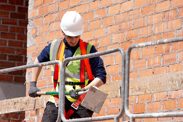 Worker in a safety helmet and safety equipment works with a tool against the background of a brick wall near protective barriers at a height.  Building Construction Process