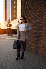 Chic woman in a plaid skirt and sunglasses by a brick wall.