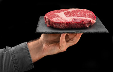 Cook holds graphite serving board with raw ribeye steak on black background.