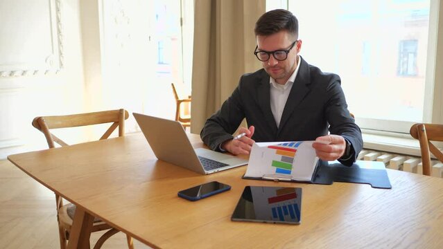 A man is a manager in the office, looks at the report on the documents for the month and enters it into an online table on a laptop.