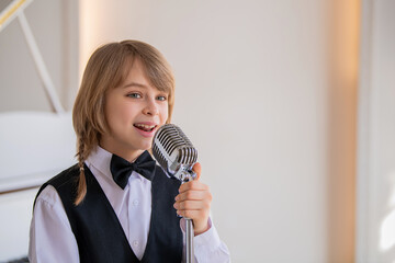 A boy in a black vest and bow tie sings into a microphone.
