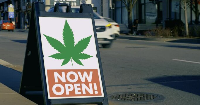 A sidewalk sign for a marijuana dispensary business in the downtown area of a large city. Traffic passes in the background.	