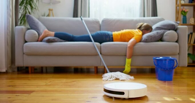 Person lying on sofa with mop, robotic vacuum cleaner on hardwood floor.