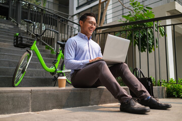Smiling businessman using laptop on staircase in the city near his bicycle.