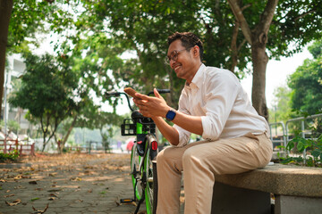 Happy adult businessman sitting on a bench next to his bicycle in city park and using his mobile...