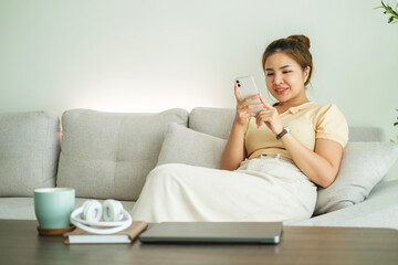 Pretty young woman in casual clothes using mobile phone, sitting on couch in cozy living room