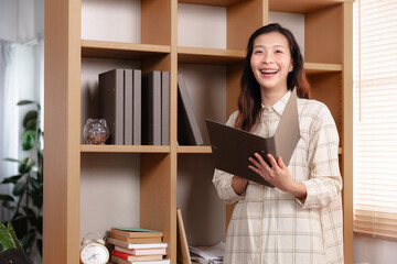 Asian freelance small business entrepreneur female start-up happy working at cozy home office with business performance, smiling businesswoman looking camera standing at desk in condominium apartment