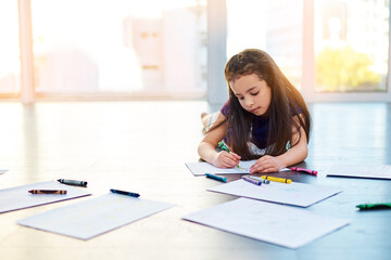 Female child, writing and drawing on floor in house with color pencil for development. Girl, playing and learning in home with crayon or paper for kindergarten, homework and homeschool education