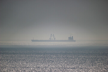 A long ship stands next to the cranes on a sea gray horizon in the fog