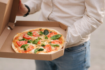 Middle selection of young man opening pizza box outdoors. Fresh vegetarian pizza, basil, tomatoes,...