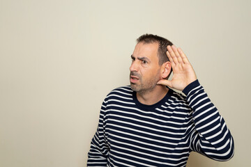Hispanic man with beard in his 40s wearing a striped sweater listening to rumors keeping hand near ear isolated over beige color background.