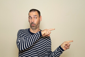 Bearded Hispanic man in his 40s wearing a striped sweater pointing to the side with his index fingers with an expression of surprise. Isolated on beige background.
