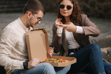 Young bloggers make photo of vegan pizza outdoor. Happy couple take food photography. Man hold...