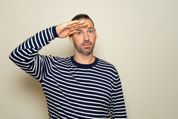 Hispanic man in his 40s saluting the camera with a military salute in an act of honor and patriotism, showing respect. Isolated on beige background.