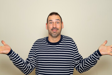 Hispanic man with beard in his 40s wearing a striped sweater clueless and confused with open arms, no idea concept. Isolated on beige studio background.