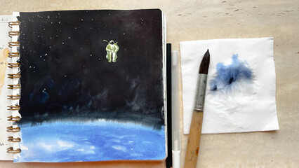 Top view of watercolor sketchbook with hand drawn illustration of cosmos and spaceman above the Earth. Aquarelle sketch on paper. Astronaut. Film grain texture. Soft focus. Blur