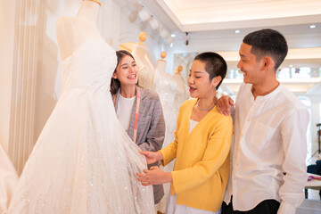 Fototapeta premium Asian bride is smiling while choosing wedding dress in modern wedding salon, Attractive designer girl using tape meter fitting on wedding dress at wedding studio.