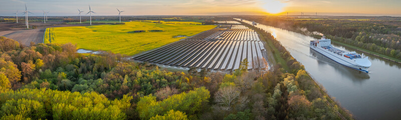 Ground-mounted solar photovoltaic panels, wind turbine farm neighboring canal, where ship...