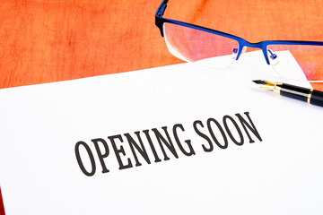 An opening soon sign an inscription on a white sheet on the table near glasses and a fountain pen