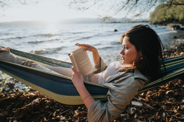 Carefree young girl deeply absorbed in reading a book, relaxing in a hammock by the serene lake as...