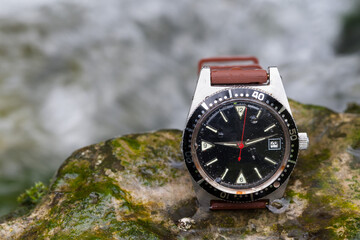 A used, sturdy outdoor watch lies on a rock by the rushing stream, embodying unwavering reliability...