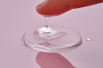 A finger touches a drop of cosmetic product on a pink background.