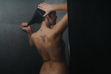 A back view of a woman washing her hair under a shower, water drops and a tattoo visible.
