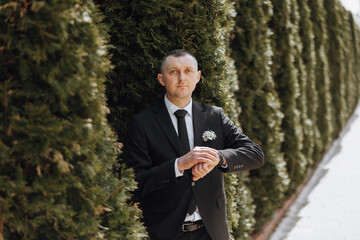 A man in a suit stands in front of a hedge, wearing a tie and a boutonniere - obrazy, fototapety, plakaty