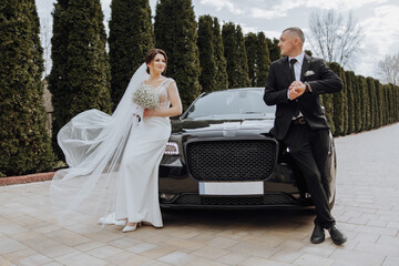 the bride and groom pose near a black car in nature