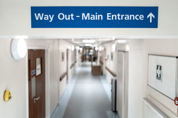 Shallow focus of a Way Out and Main Entrance sign seen within the corridor of an NHS hospital in...