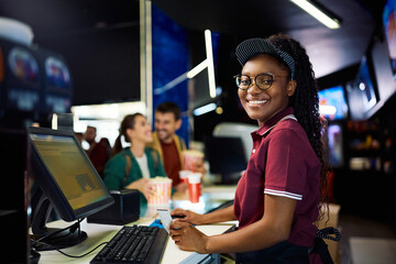 Happy black female cashier using desktop PC while working in movie theater and looking at camera.