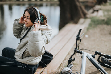 A young woman sits by the water in a park listening to music on headphones, with her bicycle beside...