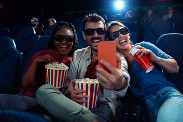 Cheerful friends taking selfie during 3D film projection in movie theater.