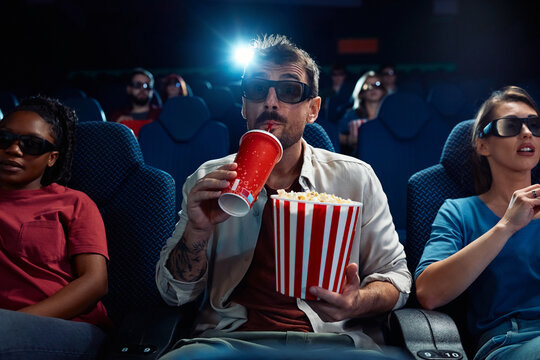 Young man drinking soda and eating popcorn while watching 3D movie in theater.