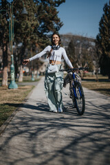 Naklejka premium Joyful young woman with headphones walking alongside her bike on a sunny park path, exuding happiness and an active lifestyle.