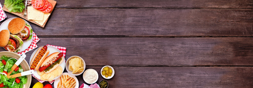 Summer BBQ or picnic corner border with hamburgers, hotdogs, salad and snacks. Top down view over a dark wood banner background. Copy space.