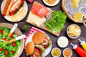 Summer BBQ or picnic table scene with hamburgers, hotdogs, salad and snacks. Overhead view on a dark wood background. - obrazy, fototapety, plakaty