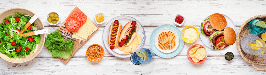 Summer BBQ or picnic table scene with hamburgers, hotdogs, salad and snacks. Above view over a white wood banner background.