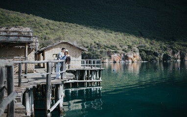 A joyful couple stands on an old wooden dock, surrounded by serene water and hillside, embodying a...