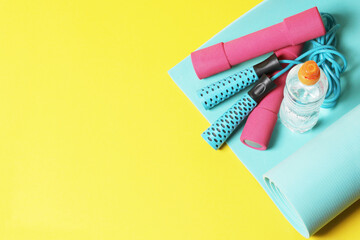 Fitness items jump rope, dumbbells, bottle of water and exercise mat on a yellow background