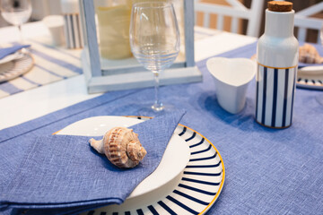 A table with a blue and white striped tablecloth and a shell on a napkin. The table is set for a...