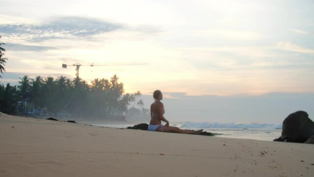 Woman in bikini engages in yoga session on sandy beach near ocean. Lady practices sitting straddle asana and mist covers warm water